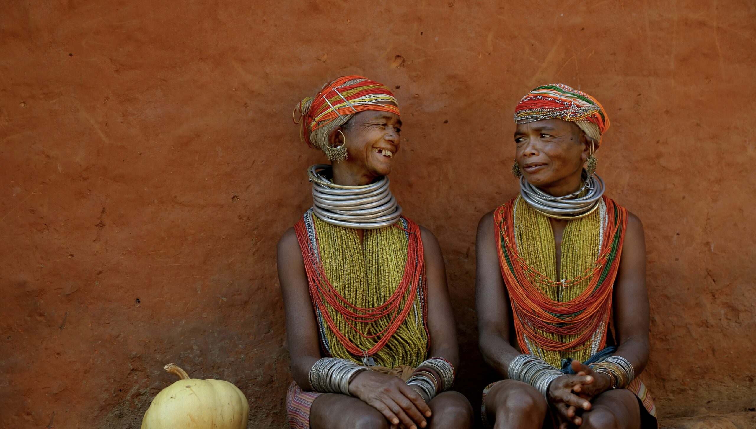 Two women wearing colorful traditional jewellery and smiling in front of a clay wall